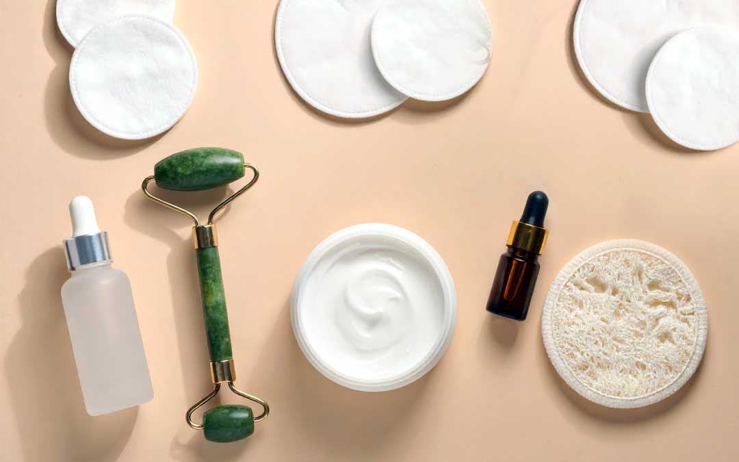 Skincare routine essentials flat lay with jade roller serum cream and cotton pads on a beige background.